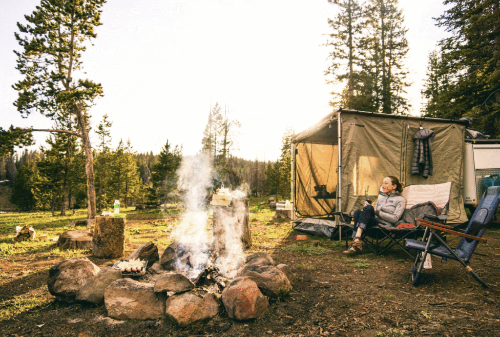 woman-lounging-on-camping-chair-in-front-of-bonfire-in-sunny-forest-with-square-tent-behind-her